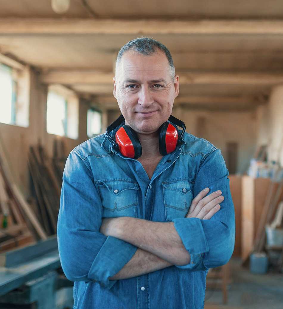 A carpender standing with crossed arms smiling while waering a ear protector around his neck