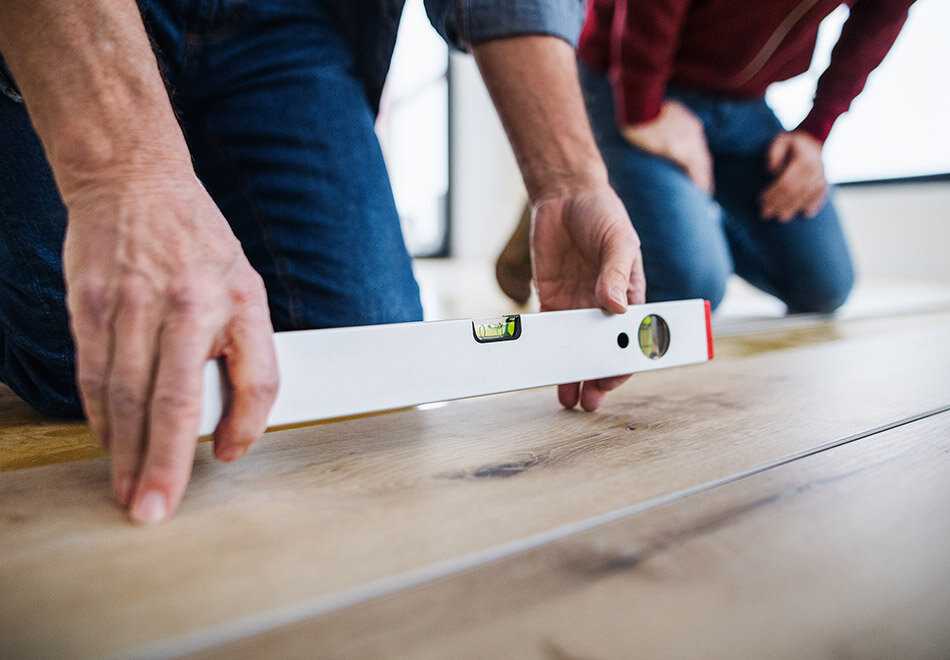 Two men kneeing on a woodfloor while one man holding a leveller tool